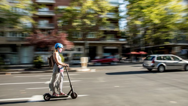 Woman riding an e-scooter down a main road, she is wearing a blue helmet. In the background trees, cars and buildings are blurred out.