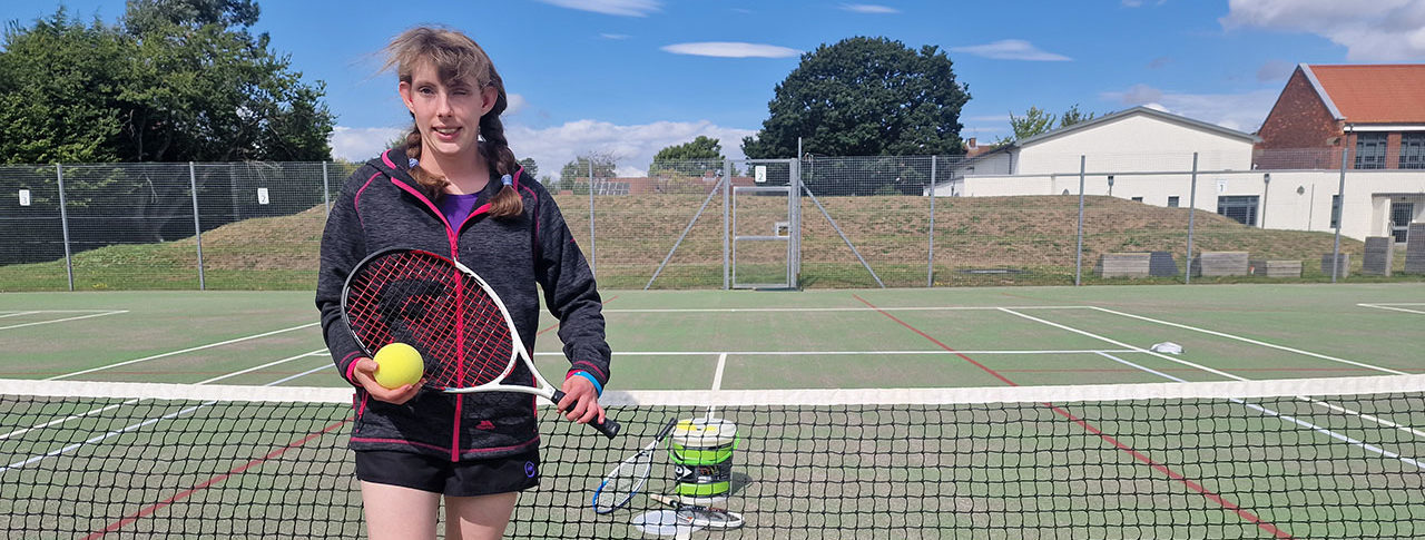 Lydia Wrightson, Ladies B4 Single Champion at the National VI Tennis Championships and a supporter coach at the event, stood in a tennis court holding a racket and ball.