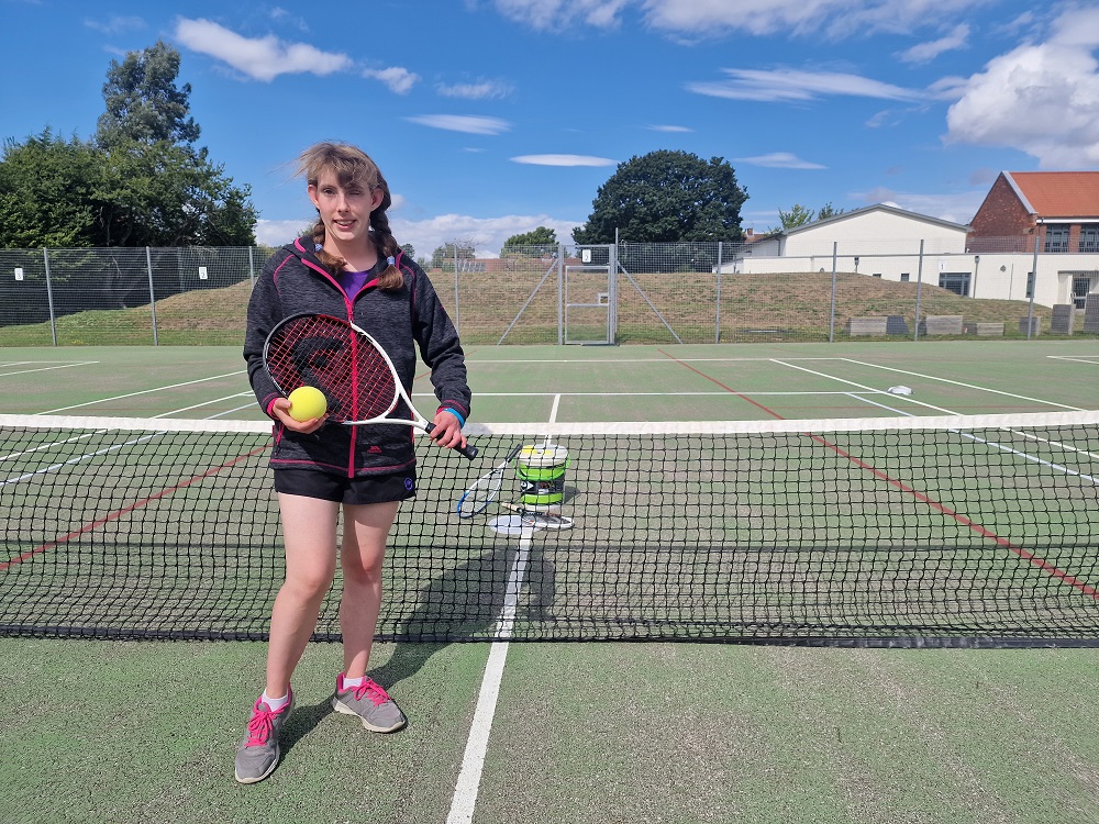 Lydia Wrightson, Ladies B4 Single Champion at the National VI Tennis Championships and a supporter coach at the event, stood in a tennis court holding a tennis ball and racket