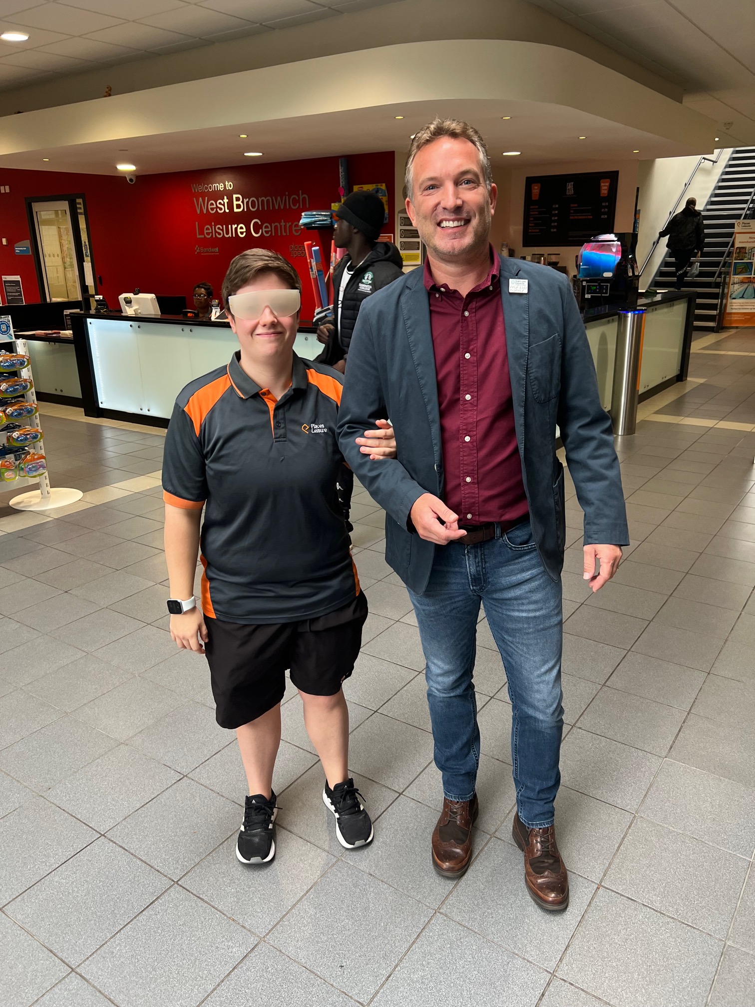 Image shows a member of staff from Places Leisure, West Bromwich, with Martyn Symcox, Head of Sport & Leisure at Thomas Pocklington Trust. The staff member is wearing sim specs, and holding on to Martyn's arm. Both are looking at the camera and smiling.
