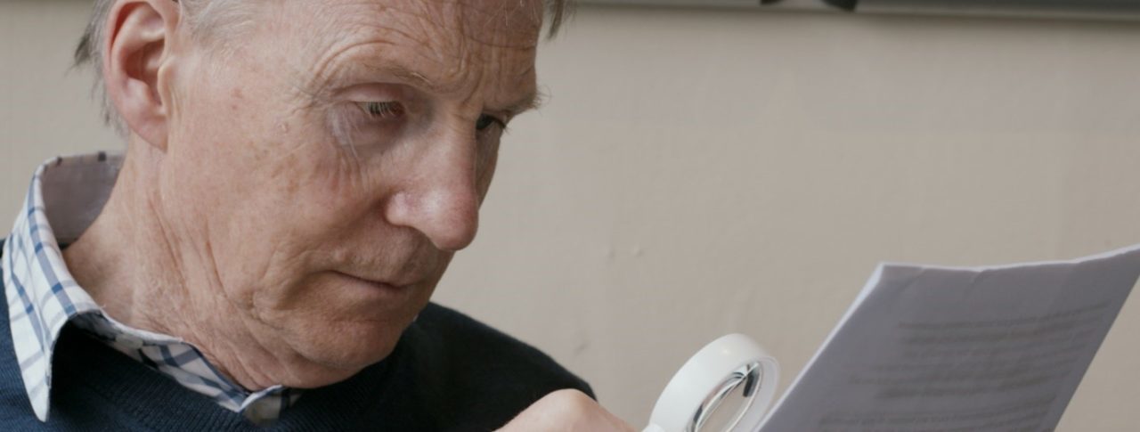 A man in a blue jumper reads from a piece of paper using a magnifier