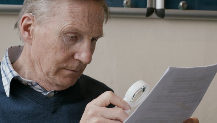 A man in a blue jumper reads from a piece of paper using a magnifier