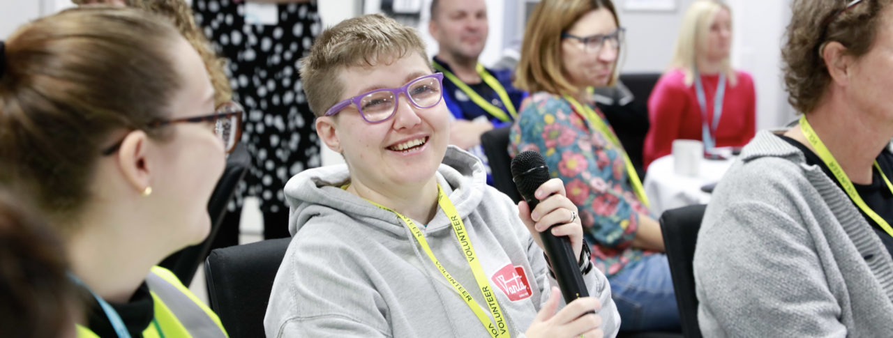 Sight Loss Council volunteer Emma, who is partially sighted, is holding a microphone ready to share her views at a volunteer event. She is sat with other delegates at a table.
