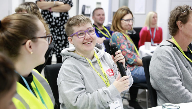 Sight Loss Council volunteer Emma, who is partially sighted, is holding a microphone ready to share her views at a volunteer event. She is sat with other delegates at a table.