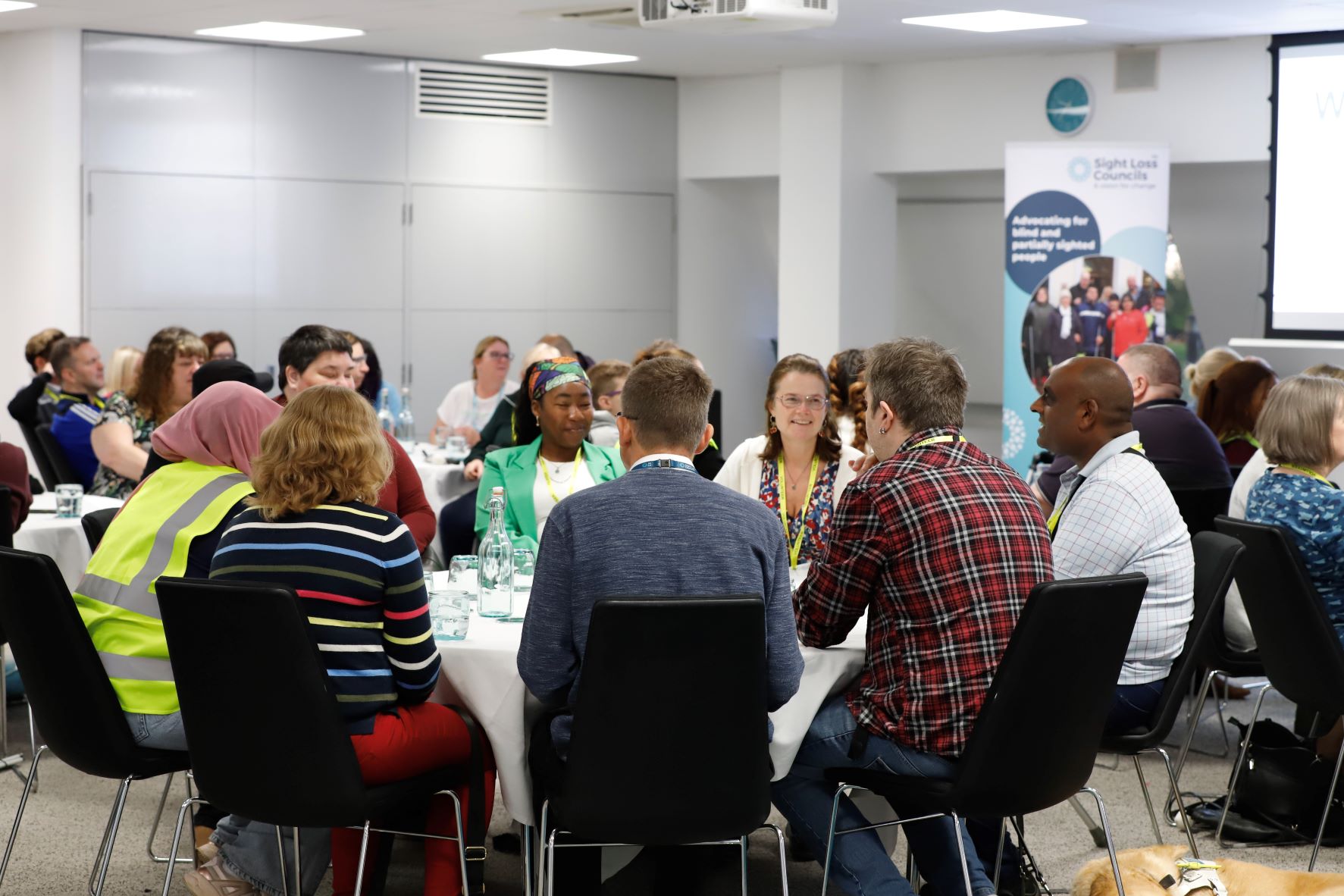 : Blind and partially sighted volunteers are sat around circular tables having a discussion at a volunteer event. A Sight Loss Council banner is in the background.