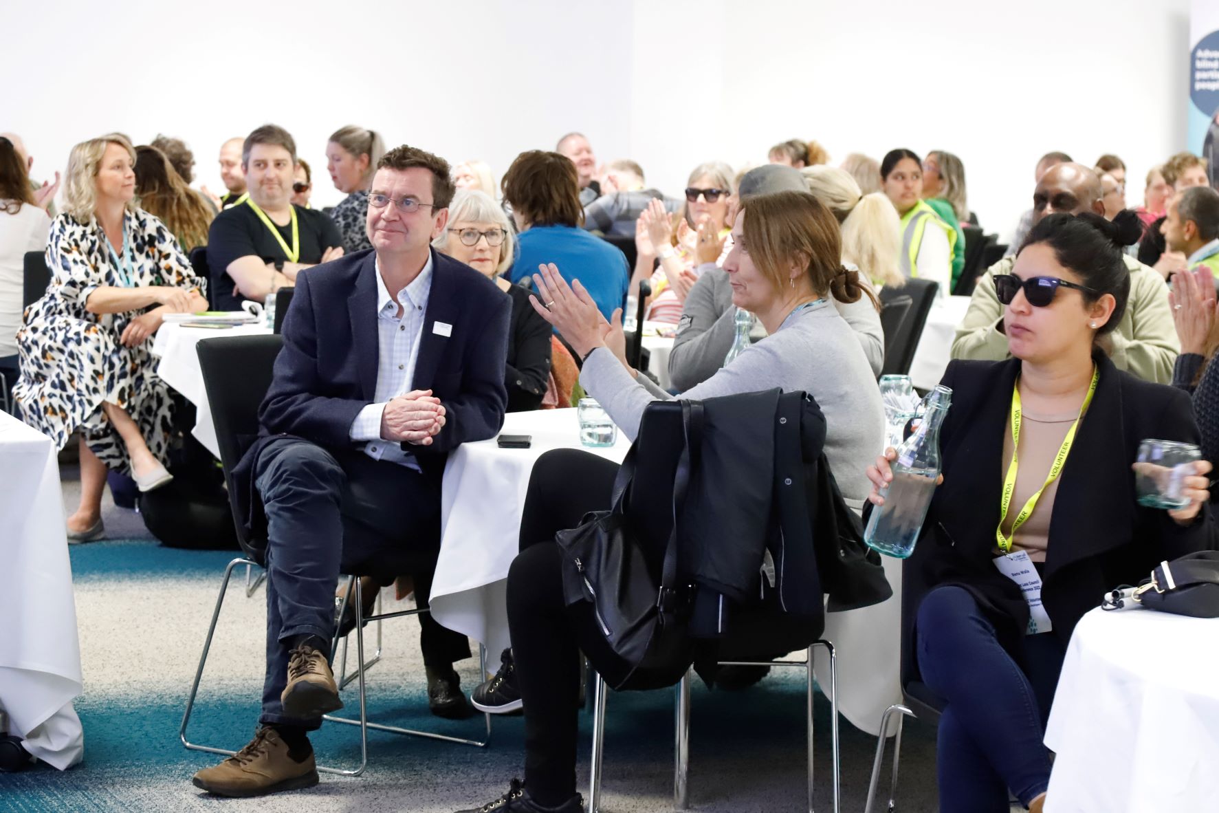 Blind and partially sighted volunteers and staff are sat around circular tables, facing forwards, listening to a presentation.