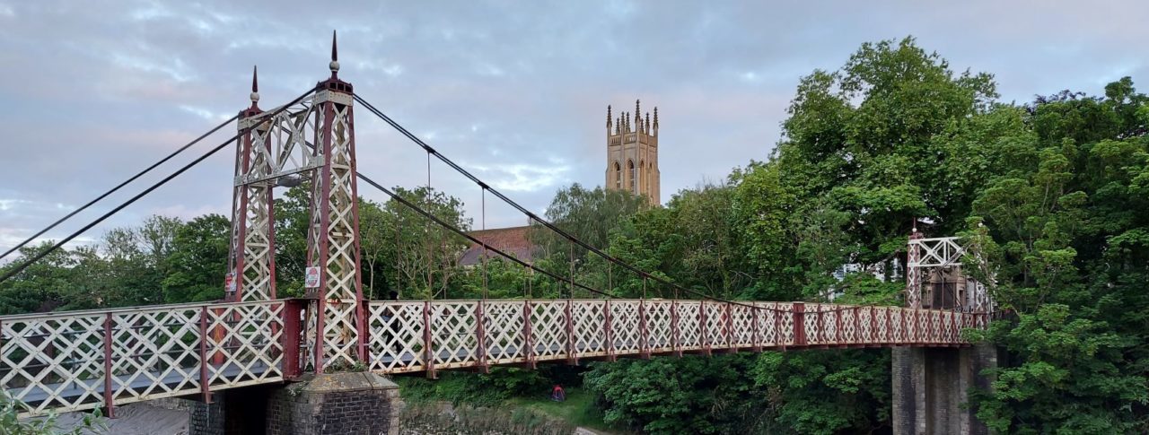 Image of Gaol Road Bridge, a colourful and ornate bridge in Bristol. There are trees surrounding the bridge, which crosses a riverbed. There is a church spire in the background of the picture.