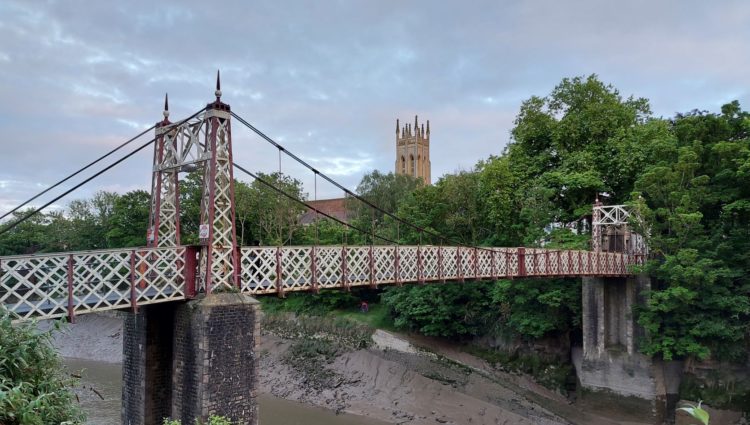 Image of Gaol Road Bridge, a colourful and ornate bridge in Bristol. There are trees surrounding the bridge, which crosses a riverbed. There is a church spire in the background of the picture.