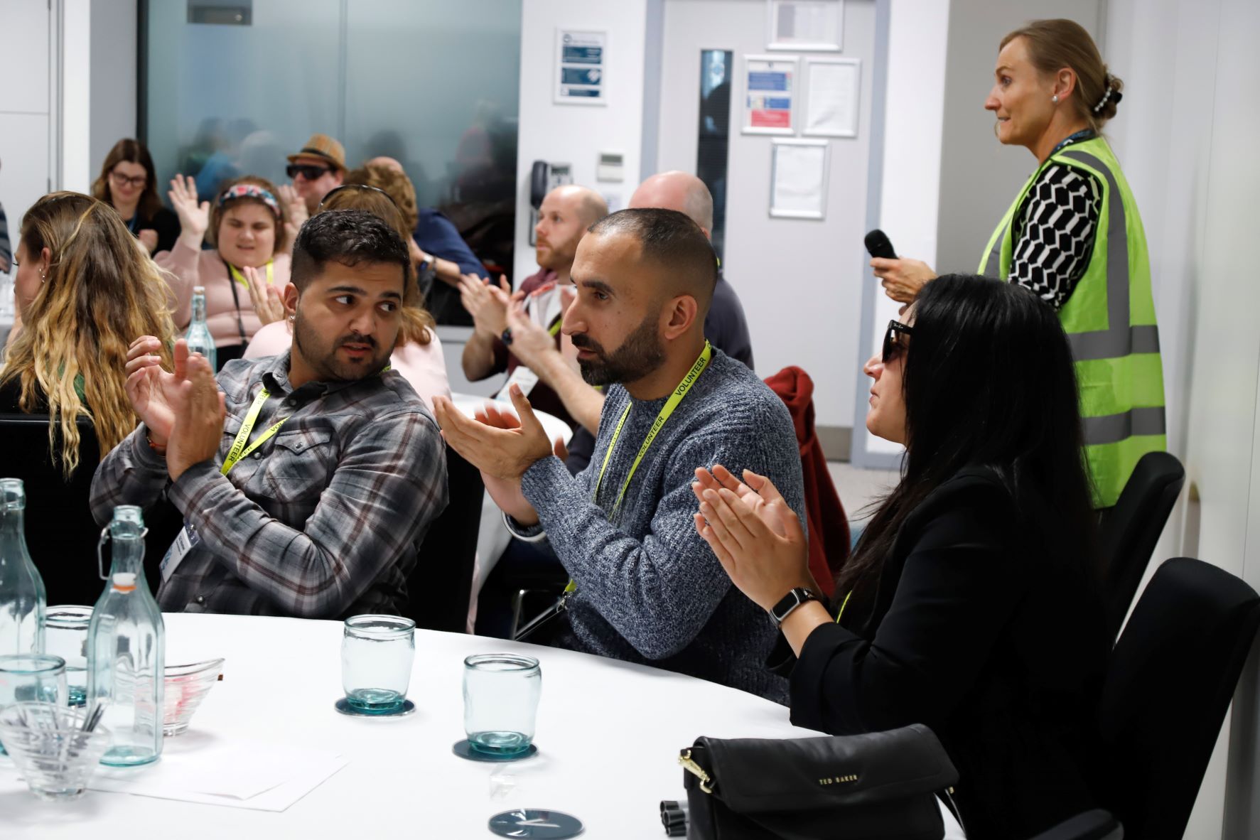 Left to right: Vinnie, support worker, is with Davinder and Renu of London SLC. They are seated at a table during a workshop, clapping. Vinnie is looking over his shoulder at Davinder.