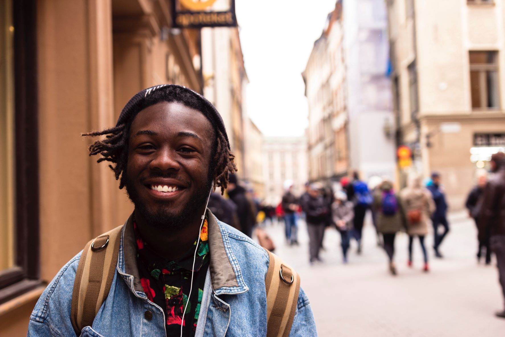 Image shows a man walking down the street, wearing headphones. He is smiling broadly. In the background, other pedestrians are seen, blurred out.