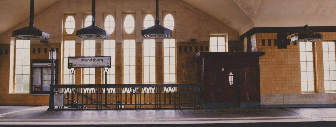 Image shows a train platform with large windows next to an old staircase. There are three art-deco lights, hanging over the platform and signage. .