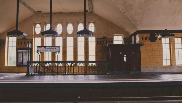 Image shows a train platform with large windows next to an old staircase. There are three art-deco lights, hanging over the platform and signage. .