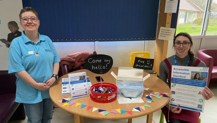 Bristol Sight Loss Council member Emma with North Bristol NHS Trust (NBT) staff member Rosie at a table with information on AIS in NBT Maternity.