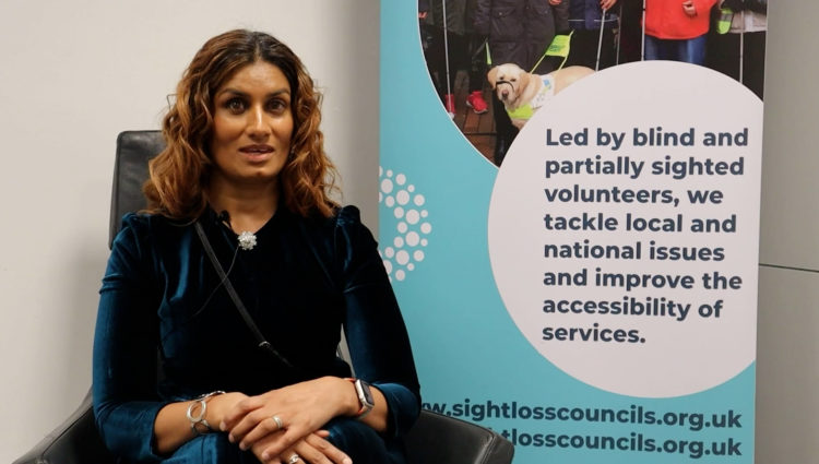 Sight Loss Council member Anela is sat on a black chair facing the camera. She has long dark hair and is wearing a green dress. There is a Sight Loss Council banner to her right. The banner says: ‘Led by blind and partially sighted volunteers, we tackle local and national issues, and improve the accessibility of services.’ It also shows the website address: www.sightlosscouncils.org.uk