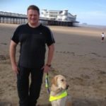 Image shows James Preston with his guide dog on a sandy beach. There is a pier in the background.