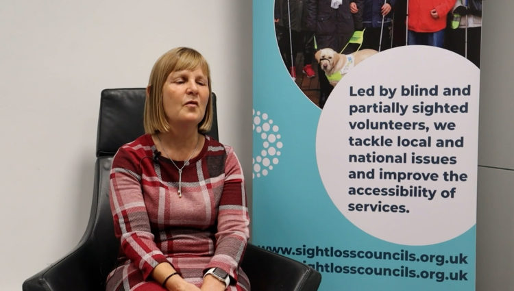 Sight Loss Council member Julie is sat on a black chair facing the camera. She is wearing a red chequered dress and has bobbed brown hair. There is a Sight Loss Council banner to her right. The banner says: ‘Led by blind and partially sighted volunteers, we tackle local and national issues, and improve the accessibility of services.’ It also shows the website address: www.sightlosscouncils.org.uk