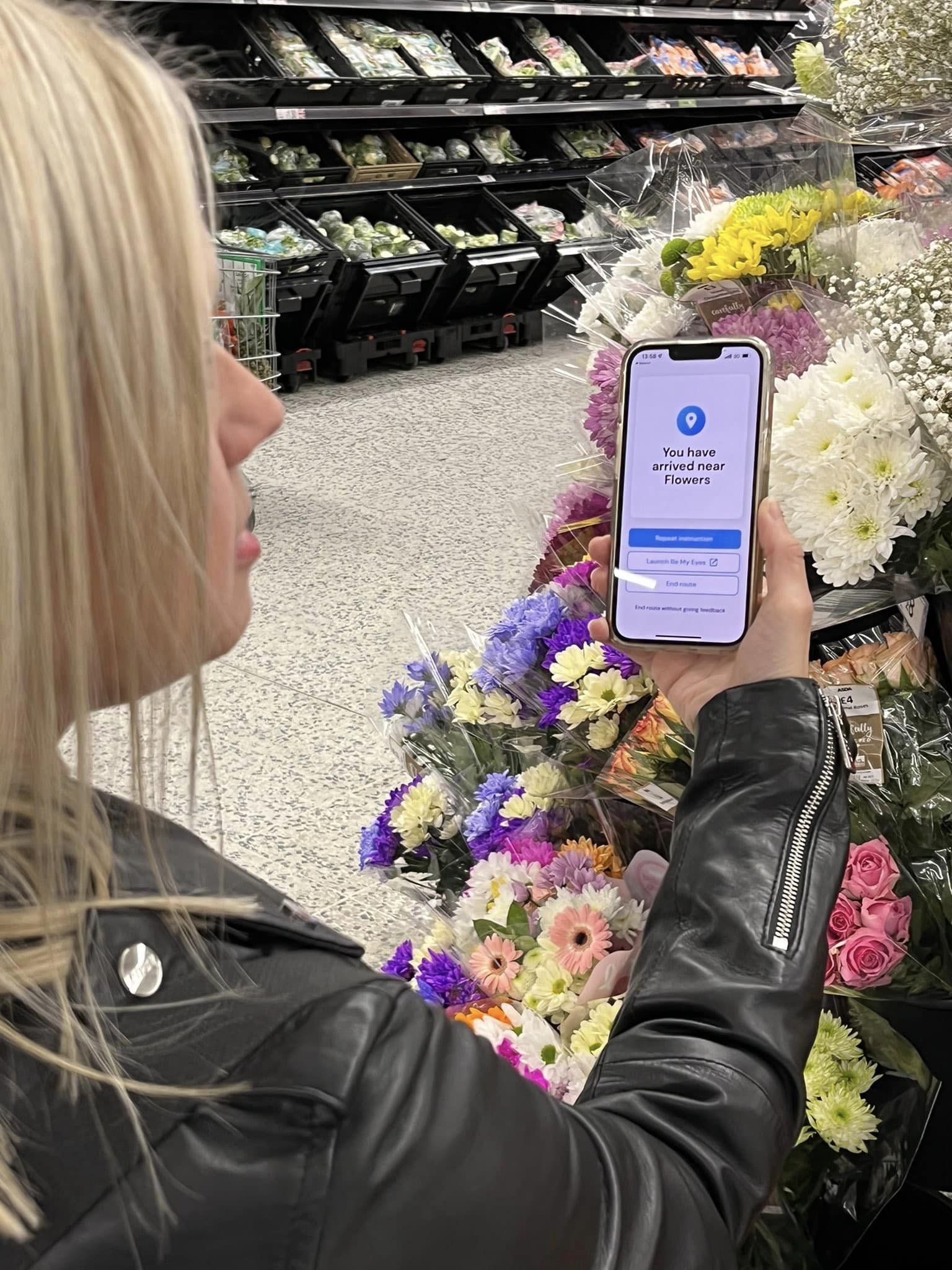 Engagement manager, Kelly Barton, is standing with her phone in her hand. She is showing the camera the GoodMaps app, which says: "You have arrived near flowers".