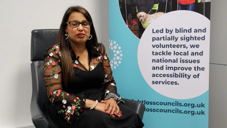 Sight Loss Council member Meena is sat on a black chair facing the camera. She is wearing a black dress with a multi-coloured flower design on the sleeves, and has long brown hair. There is a Sight Loss Council banner to her right. The banner says: ‘Led by blind and partially sighted volunteers, we tackle local and national issues, and improve the accessibility of services.’ It also shows the website address: www.sightlosscouncils.org.uk