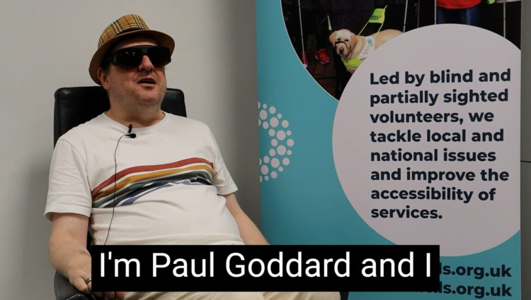 Sight Loss Council member Paul is sat on a black chair facing the camera. He is wearing a cream t-shirt with a band of multi-coloured stripes. There is a Sight Loss Council banner to his right. The banner says: ‘Led by blind and partially sighted volunteers, we tackle local and national issues, and improve the accessibility of services.’ It also shows the website address: www.sightlosscouncils.org.uk