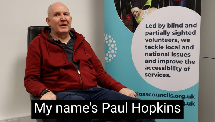 Sight Loss Council member Paul is sat on a black chair facing the camera. He is wearing a red fleece jacket and jeans . There is a Sight Loss Council banner to his right. The banner says: ‘Led by blind and partially sighted volunteers, we tackle local and national issues, and improve the accessibility of services.’ It also shows the website address: www.sightlosscouncils.org.uk