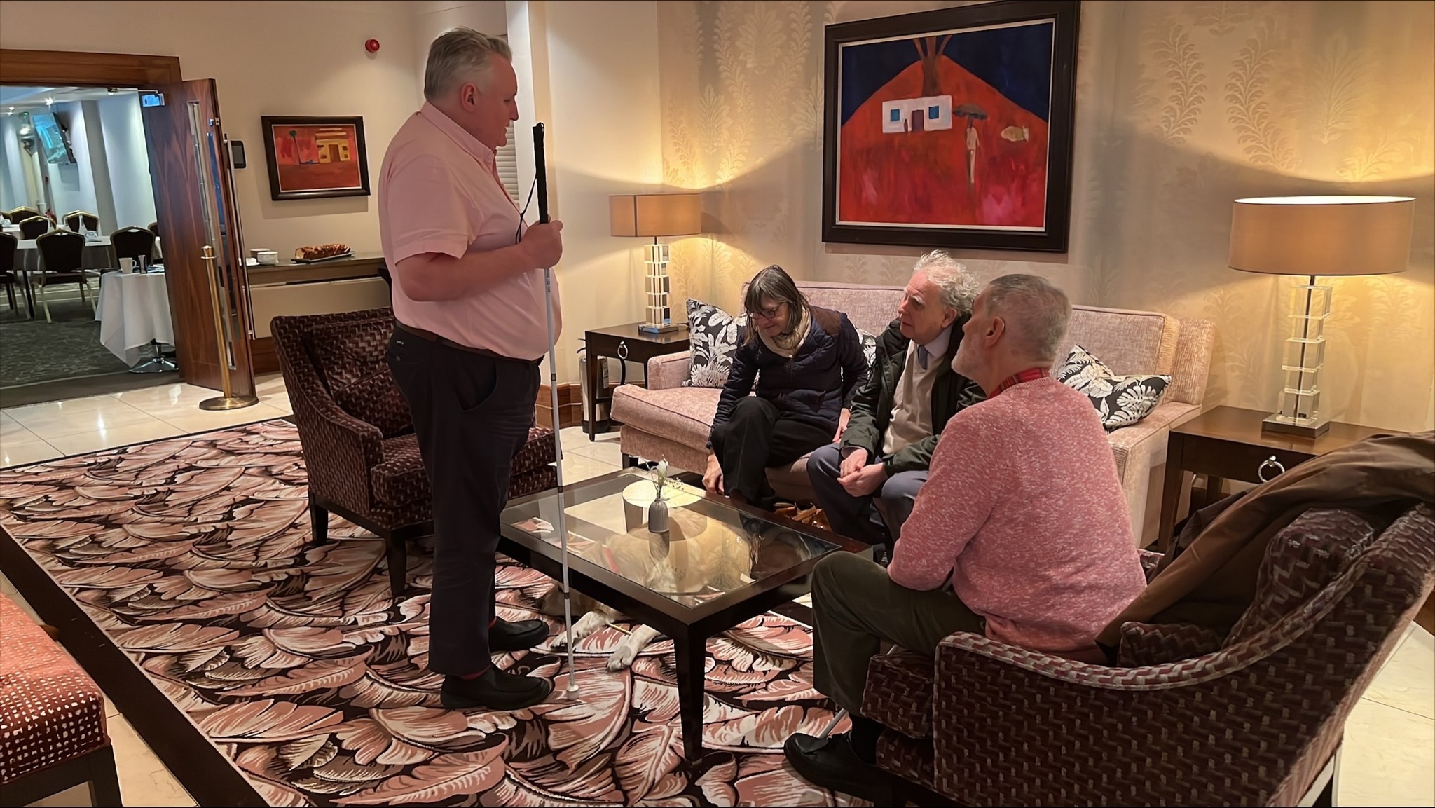 Alun Davies, Engagement Manager for the South West, talking to three attendees ahead of the cost of living VI Forum. The attendees are seated in the hotel lobby. Alun is holding his cane.