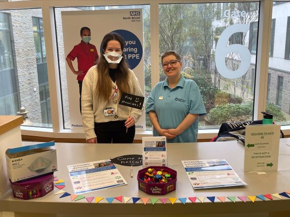 Bristol Sight Loss Council member Emma with North Bristol NHS Trust (NBT) staff member Ann who is wearing a clear face mask at a table with information on AIS in Gate 6 Brunel Building NBT.