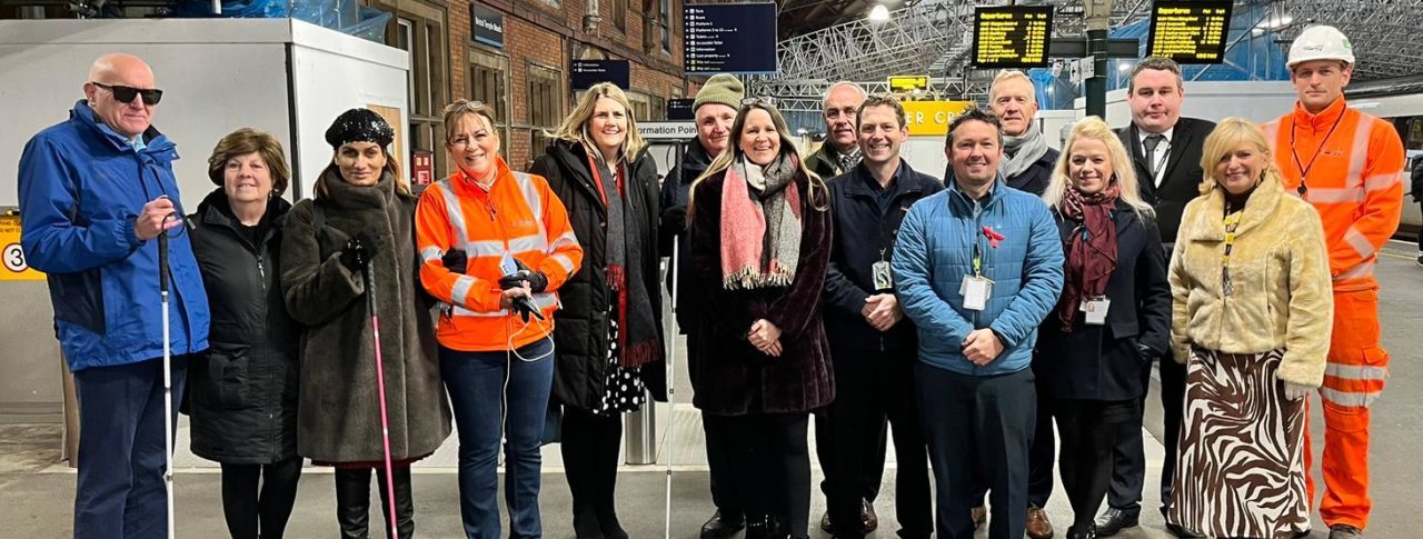 Network Rail staff and Bristol Sight Loss Council members, joined by Thomas Pocklington Trust staff, stood in a line near a platform at Bristol Temple Meads railway station. They are facing the camera and smiling.