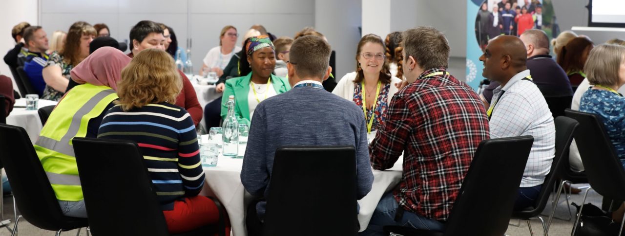 Sight Loss Council volunteers sat around a table having a discussion at a recent conference. A Sight Loss Council banner is in the background.