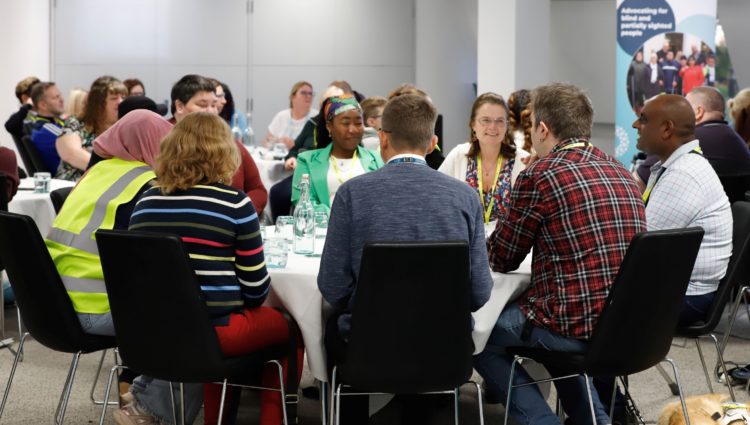 Sight Loss Council volunteers sat around a table having a discussion at a recent conference. A Sight Loss Council banner is in the background.