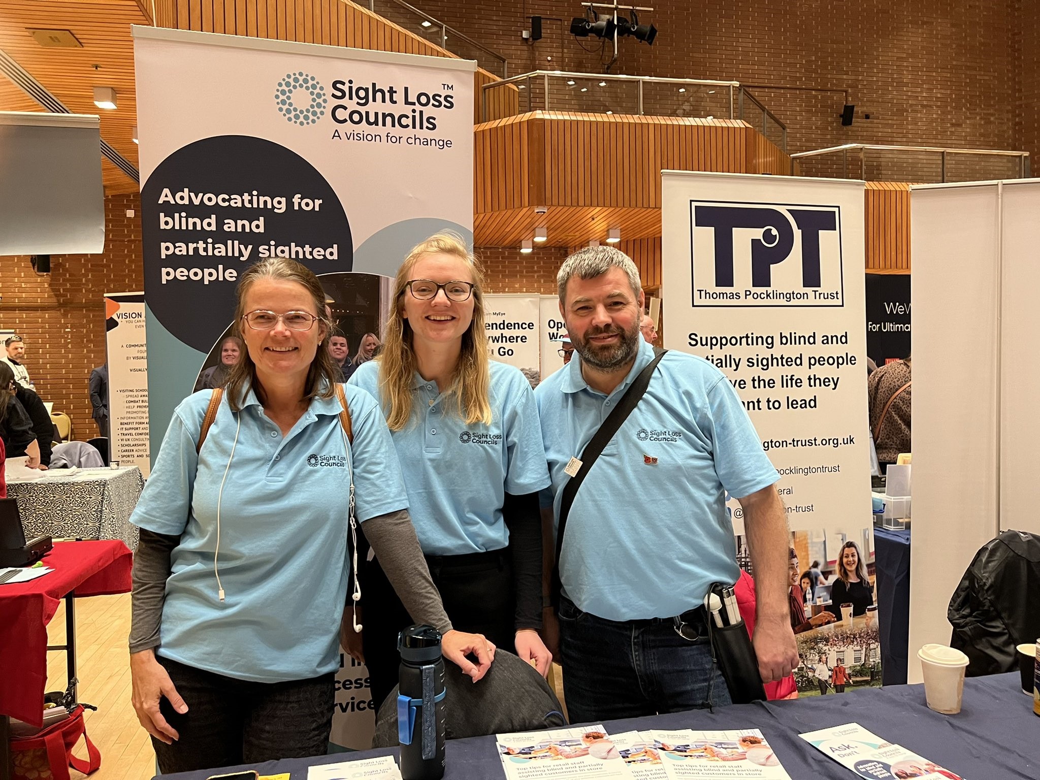 Image shows London SLC members Vicky and Steve with Senior Engagement Manager Lucy. They are behind a stall and infront of a Sight Loss Council and Thomas Pocklington Trust banner.