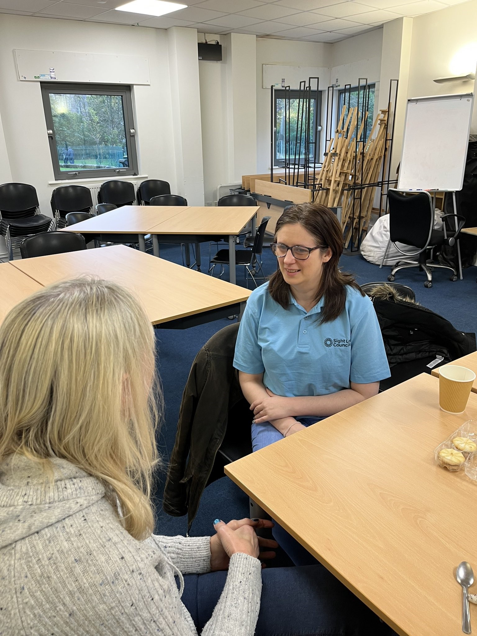 SLC member Laura listening to a member of the public at Wirral Listening Month drop-in event. They are sat down on chairs in a room surrounded by tables.