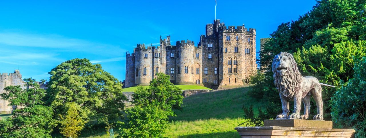 Landscape image of Alnwick Castle, Northumberland. At the forefront of the picture is a medieval stone wall, with a statue of a lion standing on it. The sky is bright blue and the image is framed by green trees.