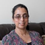 Headshot of Amrit Dhaliwal. London SLC member. Amrit is sitting on a sofa, looking at the camera, smiling. She has dark hair, pulled back. she is wearing glasses and large hoop earrings. She has a flowery top on.