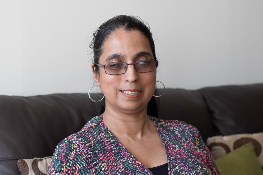 Headshot of Amrit Dhaliwal. London SLC member. Amrit is sitting on a sofa, looking at the camera, smiling. She has dark hair, pulled back. she is wearing glasses and large hoop earrings. She has a flowery top on.