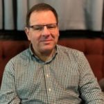Image of Graham Light, T&W Sight Loss Council member. Graham is seated. He is wearing a green and white checked shirt, wearing glasses, and smiling at the camera.