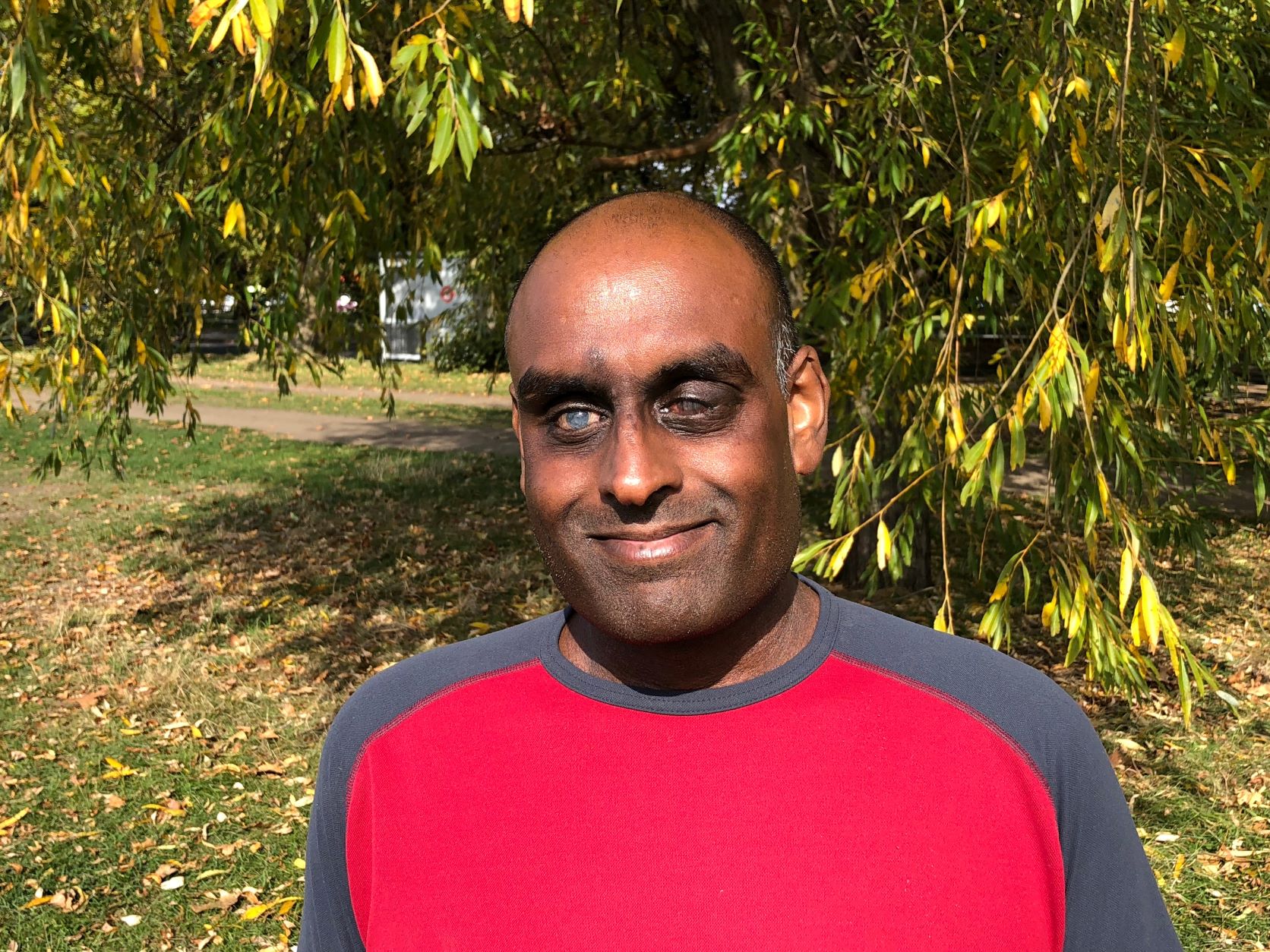 Headshot of London SLC member, Haren Thillainathan. He standing outside under a tree in the sunshine. He is wearing a red and navy t-shirt.