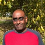 Headshot of London SLC member, Haren Thillainathan. He standing outside under a tree in the sunshine. He is wearing a red and navy t-shirt.