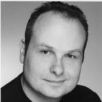 Black and white headshot of Liam O'Carroll. His head is turned slightly to the side and he is smiling.