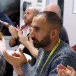 Left to right: Vinnie, support worker, is with Davinder and Renu of London SLC. They are seated at a table during a workshop, clapping. Vinnie is looking over his shoulder at Davinder.