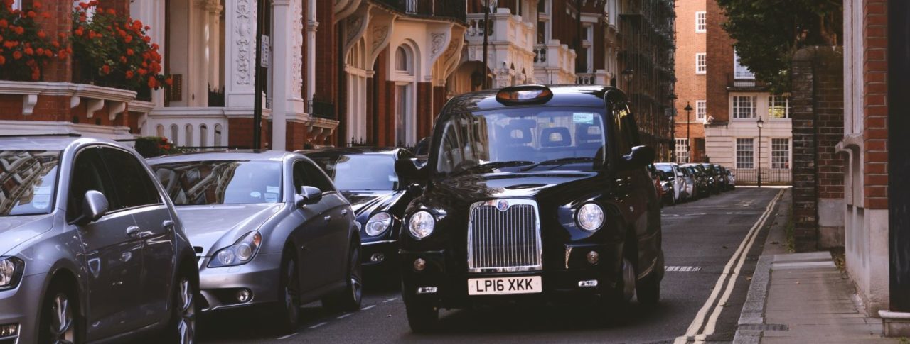 Image shows a black cab driving down a residential street in south-west London alongside a row of tall, grand, Victorian houses.