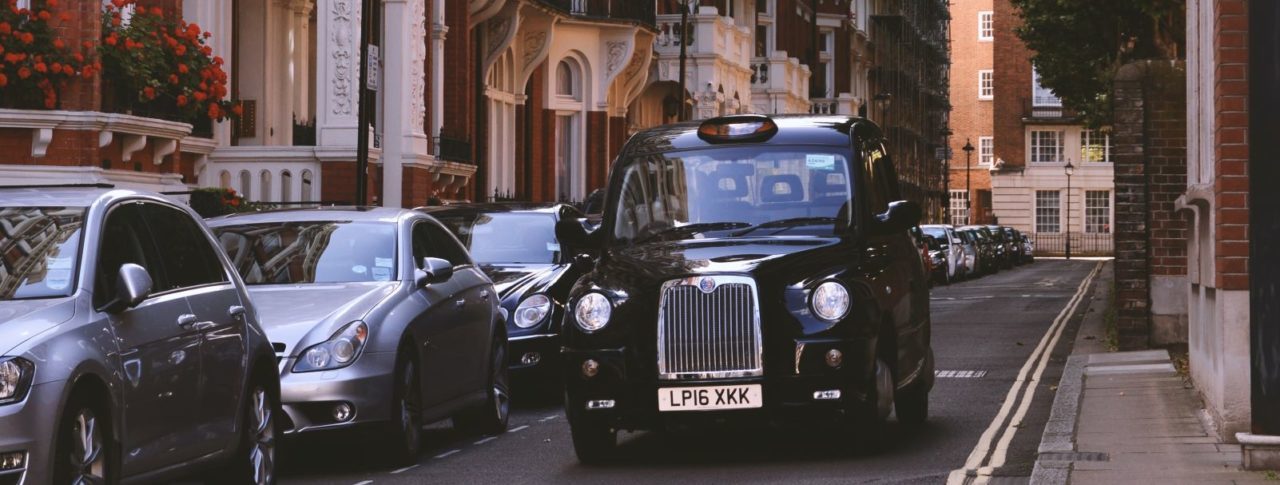 Image shows a black cab driving down a residential street in south-west London alongside a row of tall, grand, Victorian houses.