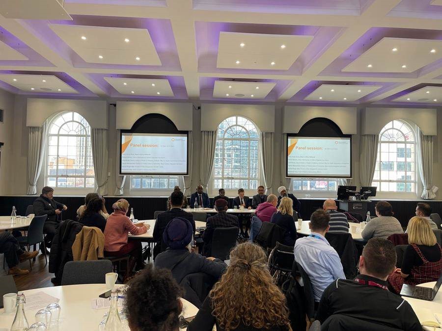 Image shows delegates sitting at round tables at the mayoral round table event, looking forward to a long table of speakers. There are projection screens either side of the speakers table.