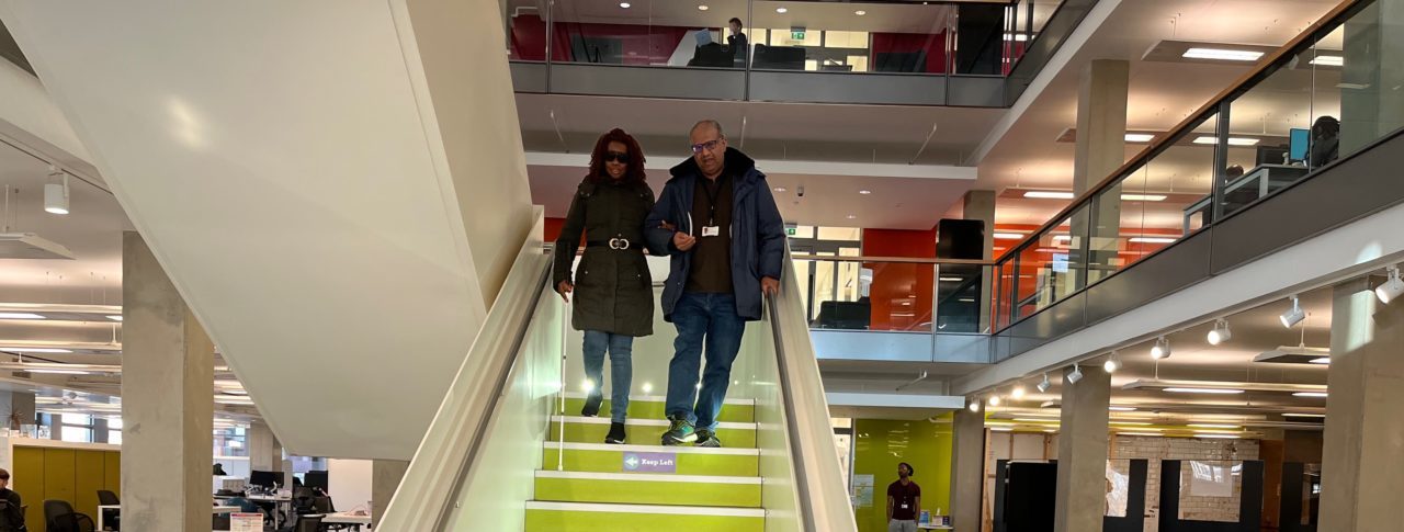 Image shows two people at the top of a staircase. The lady is holding the man's elbow. She is using a white cane, he is guiding her down the stairs.