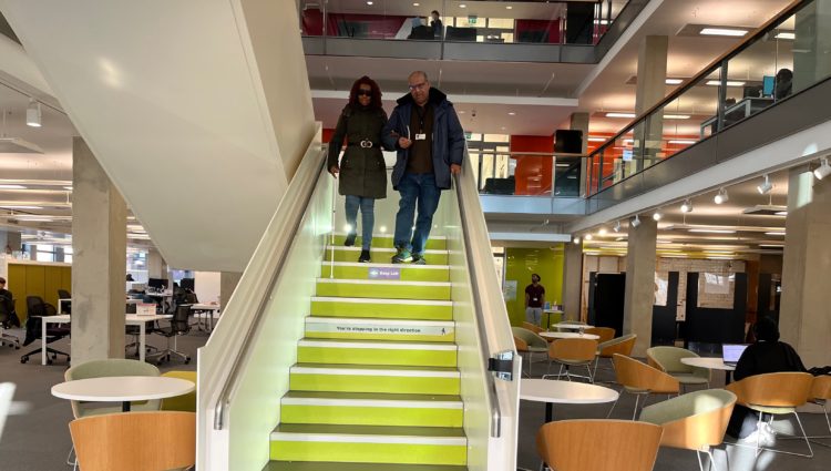 Image shows two people at the top of a staircase. The lady is holding the man's elbow. She is using a white cane, he is guiding her down the stairs.