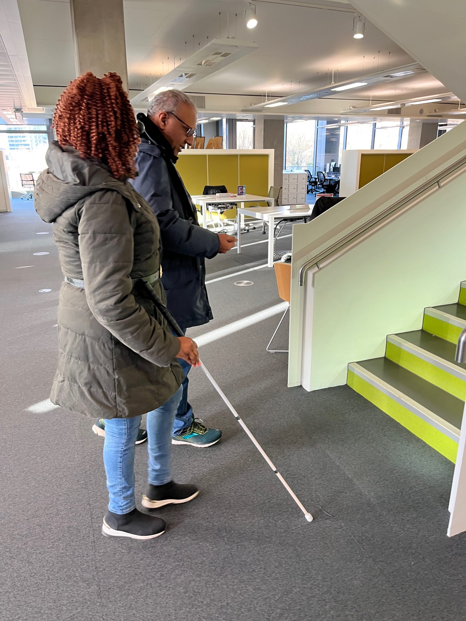 Image shows a lady holding a white cane, standing next to a man. He is about to guide her up a staircase.
