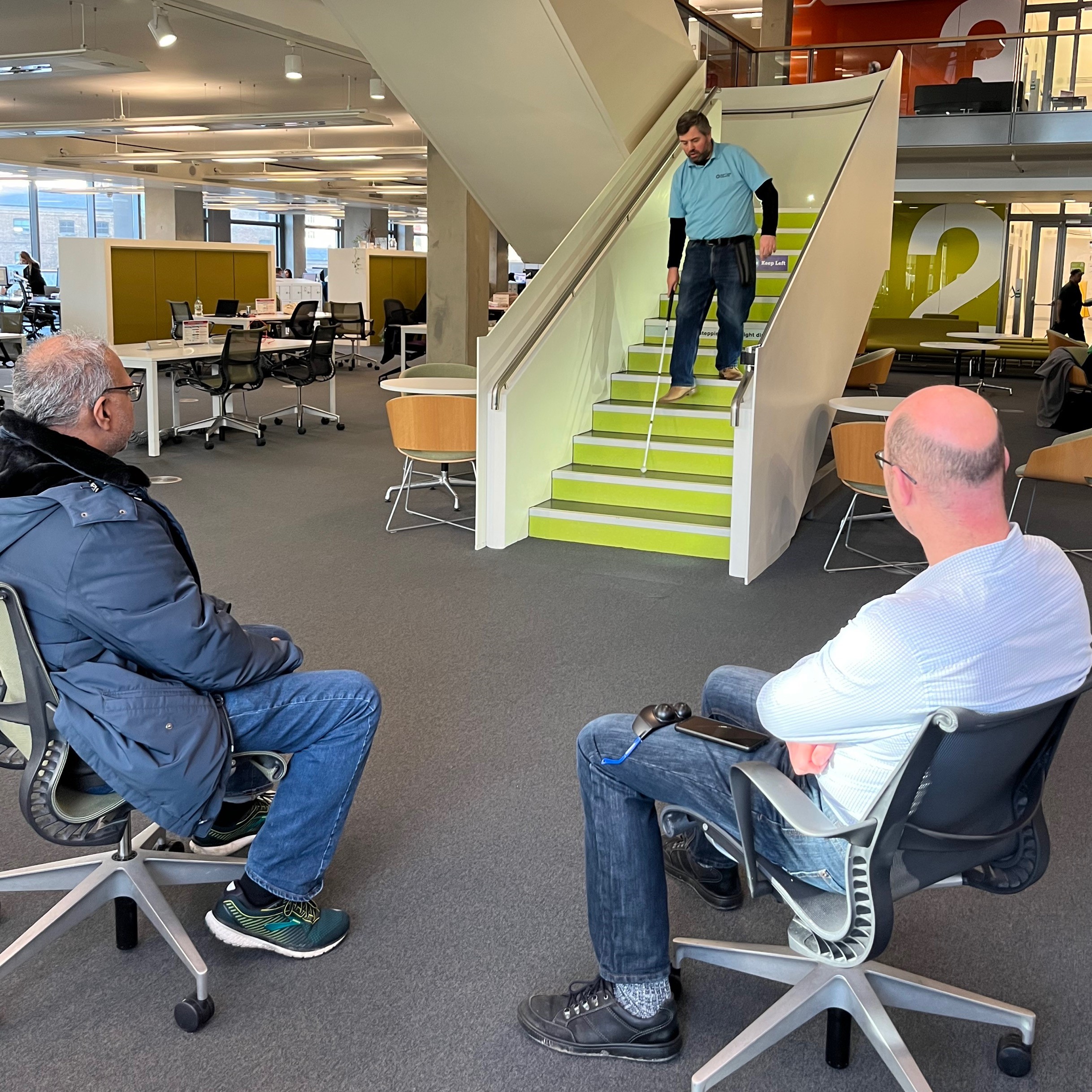 Image shows Steve Reed demonstrating walking down the stairs with his cane. Two members of the Transport Design Team are sitting at the bottom, watching him.