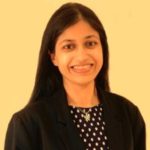 Headshot of Vidya Nathan, standing against a yellow background. Vidya has long, dark hair. She is wearing a black blazer and a polka dot top. She is smiling at the camera.
