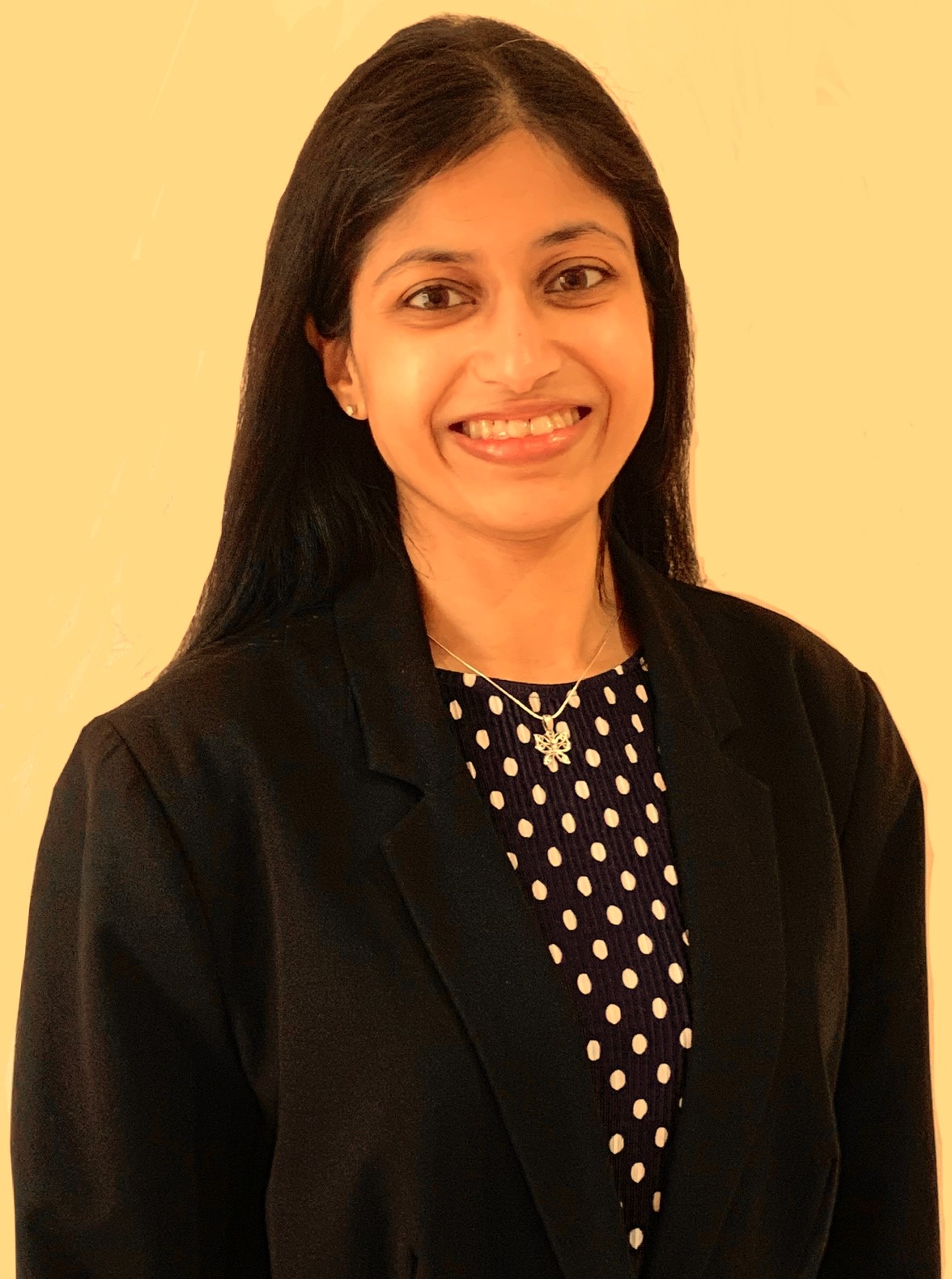 Headshot of Vidya Nathan, standing against a yellow background. Vidya has long, dark hair. She is wearing a black blazer and a polka dot top. She is smiling at the camera.