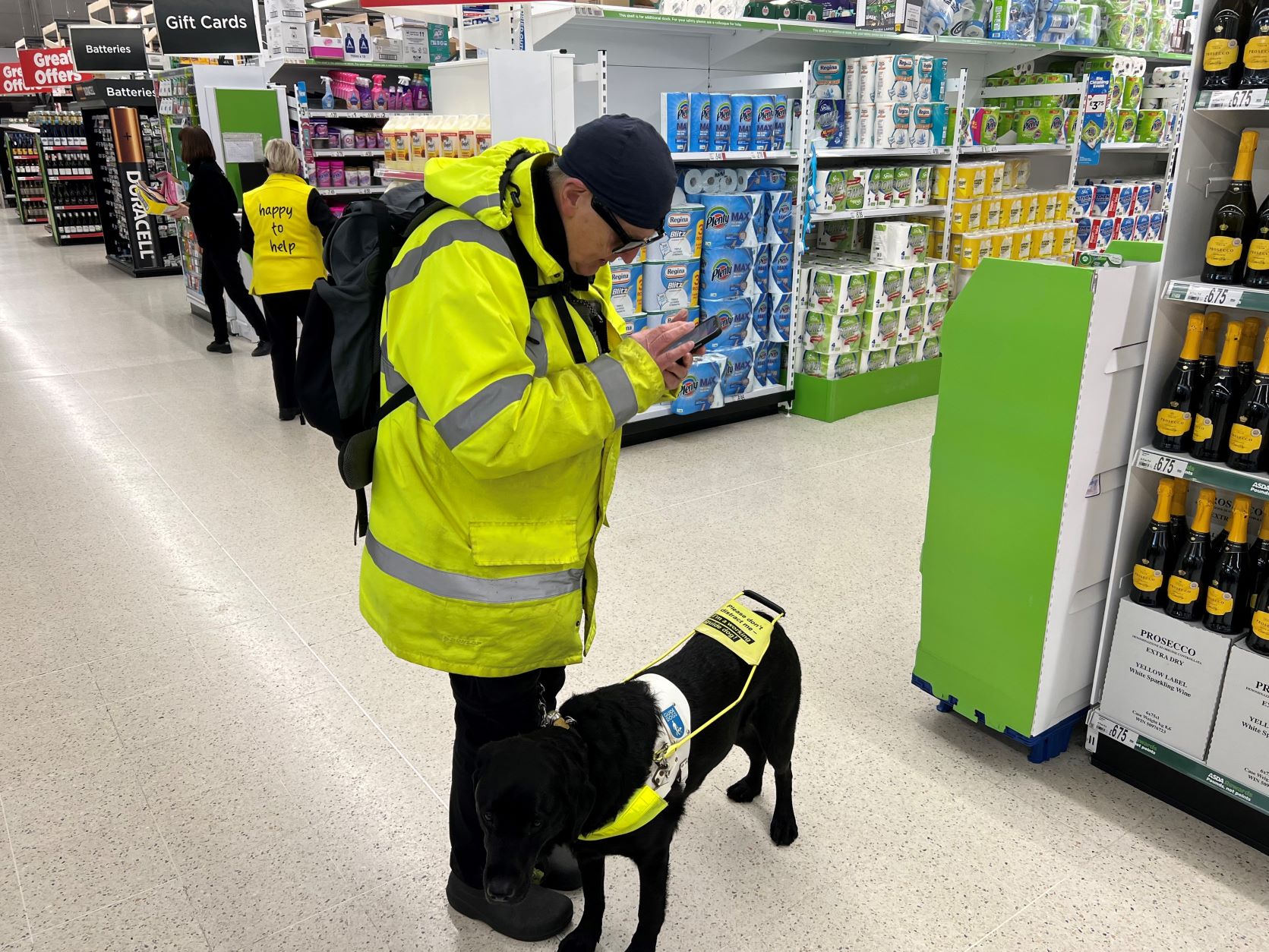Image shows David Quarmby, West Yorkshire SLC member, in an ASDA store. He is with his guide dog Reg. He is wearing a high-vis jacket, looking down at his phone whilst using the GoodMaps app.