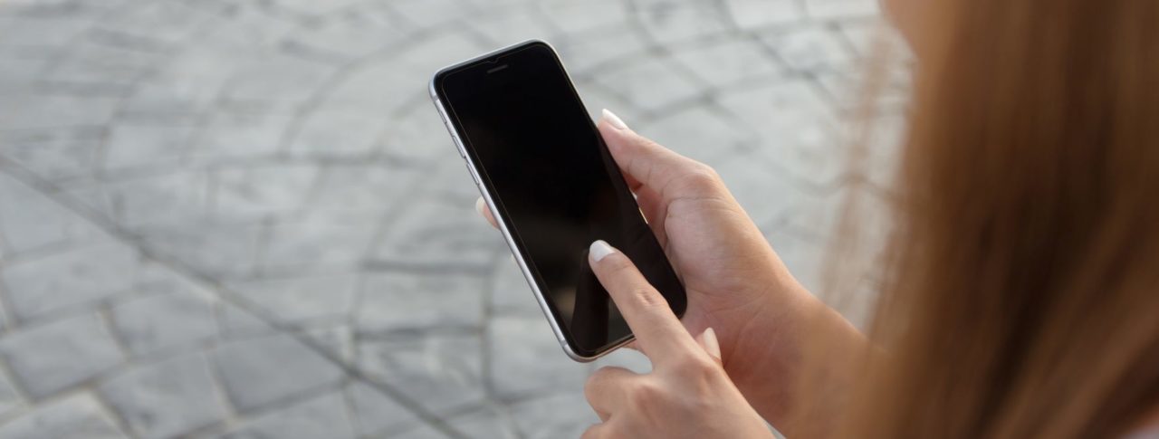 Close up on a woman's hand, holding a smartphone. Her hand finger is touching the screen. The right hand side of the image is framed by her long, red, hair.